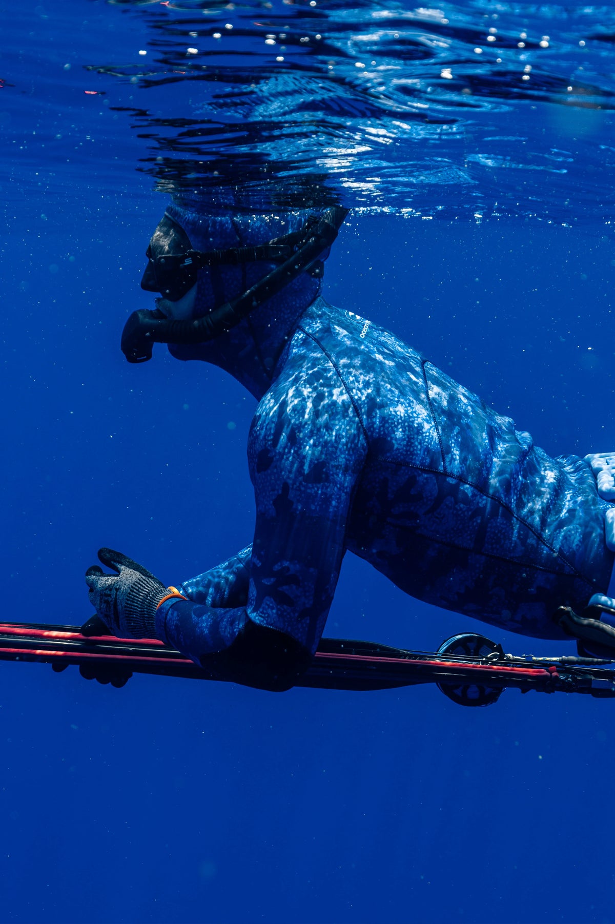 An underwater shot of a diver wearing a blue camouflage full-body wetsuit and mask, holding a spear in clear blue ocean water.