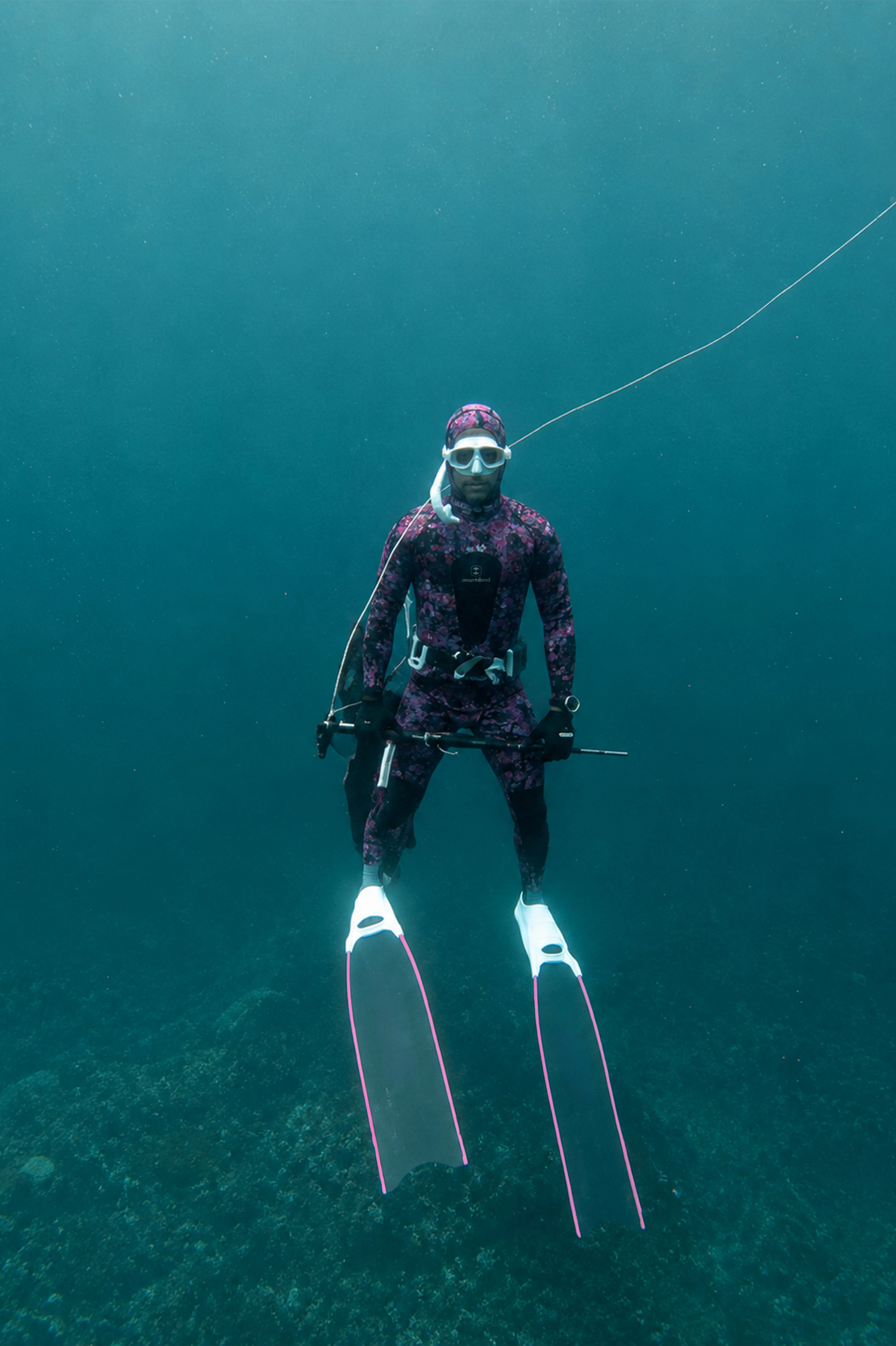 An underwater shot of a diver wearing a dark purple and black camouflage wetsuit, mask, and fins with pink trim, suspended in deep blue-green ocean water.