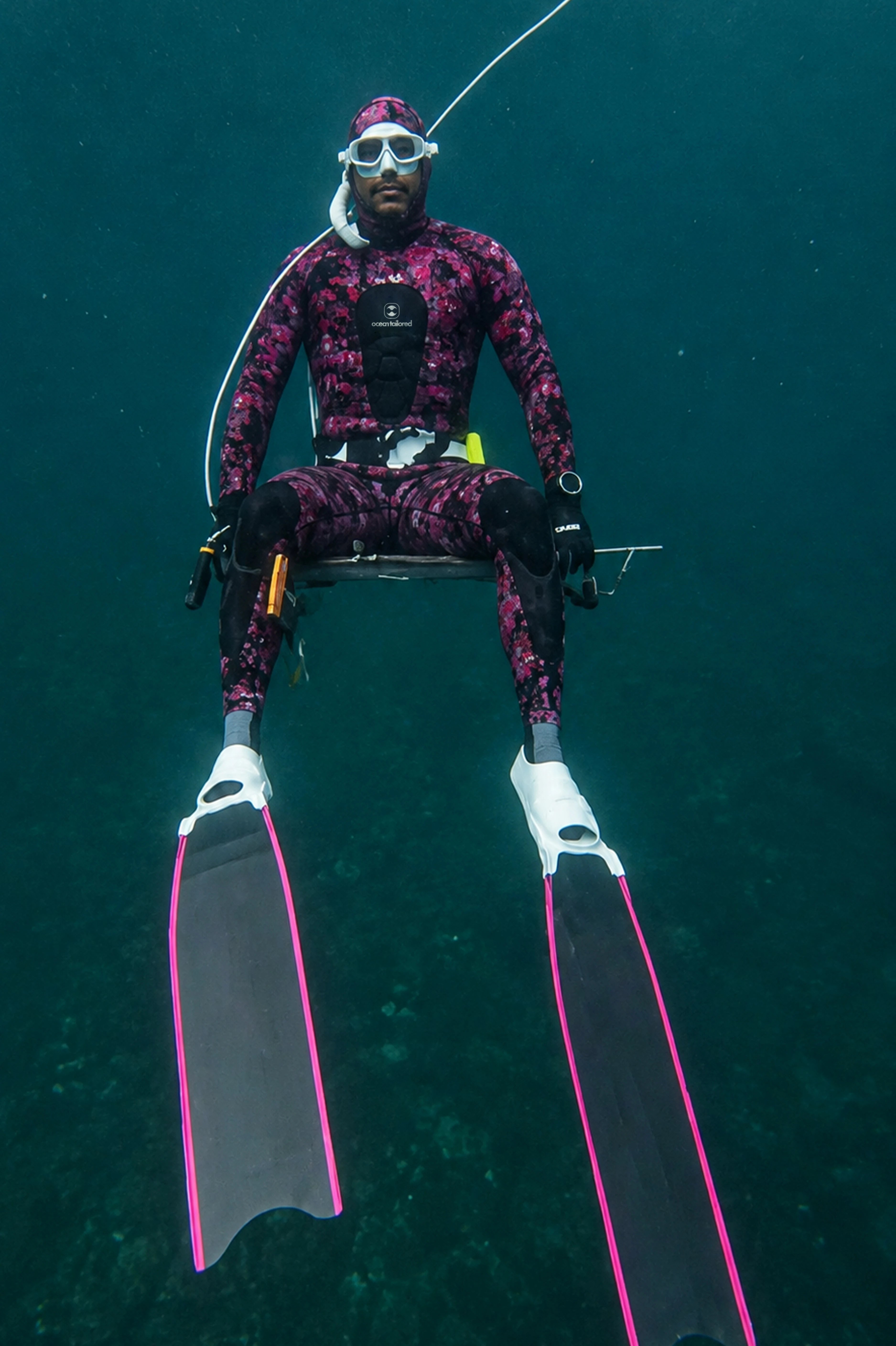 An underwater shot of a diver wearing a pink and black patterned full-body wetsuit, white mask, and long black fins with bright pink trim, suspended against deep blue-green water.