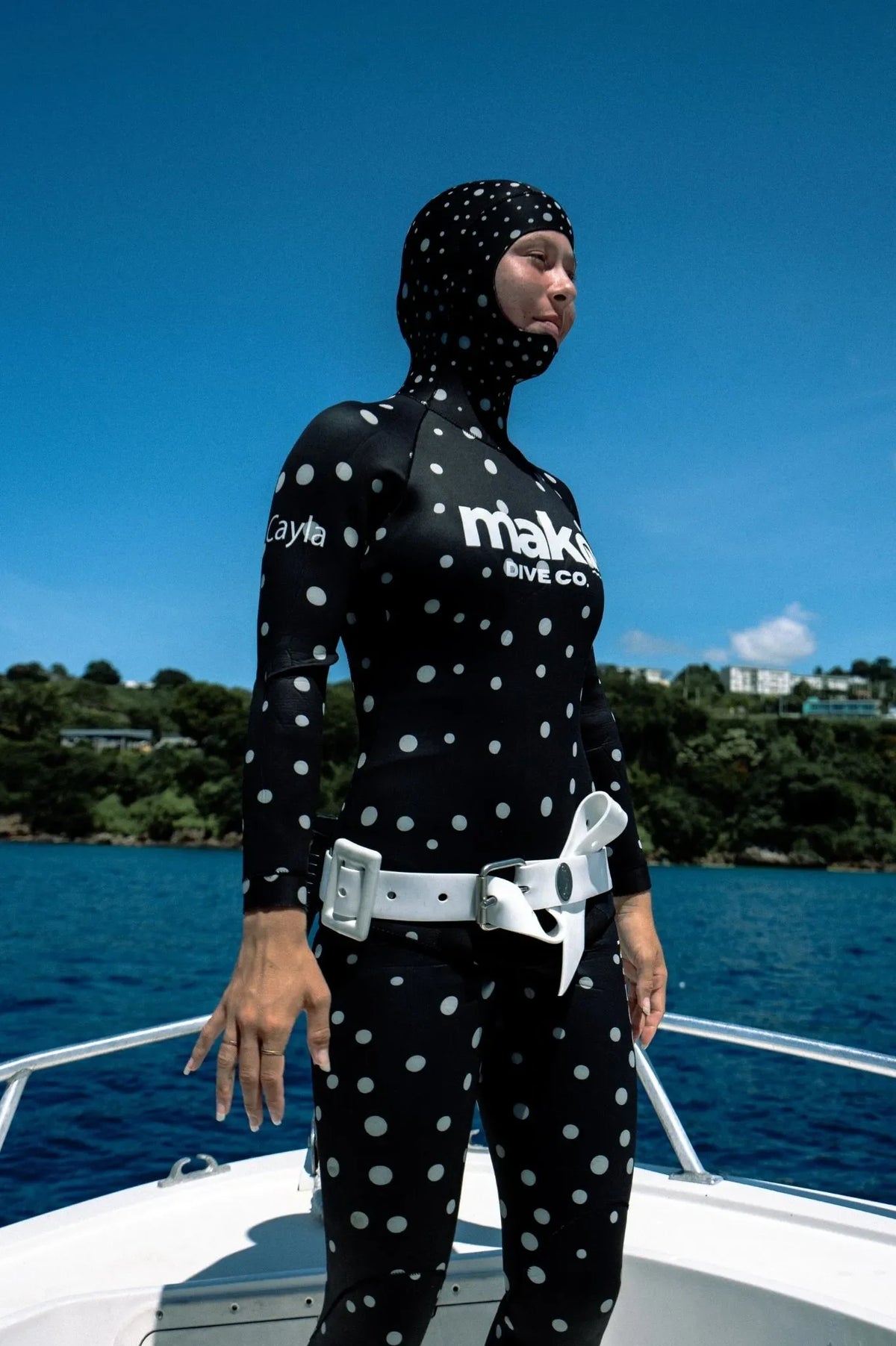Person wearing a black fully custom dream wetsuit with white polka dots and text, standing on a boat with a scenic background.