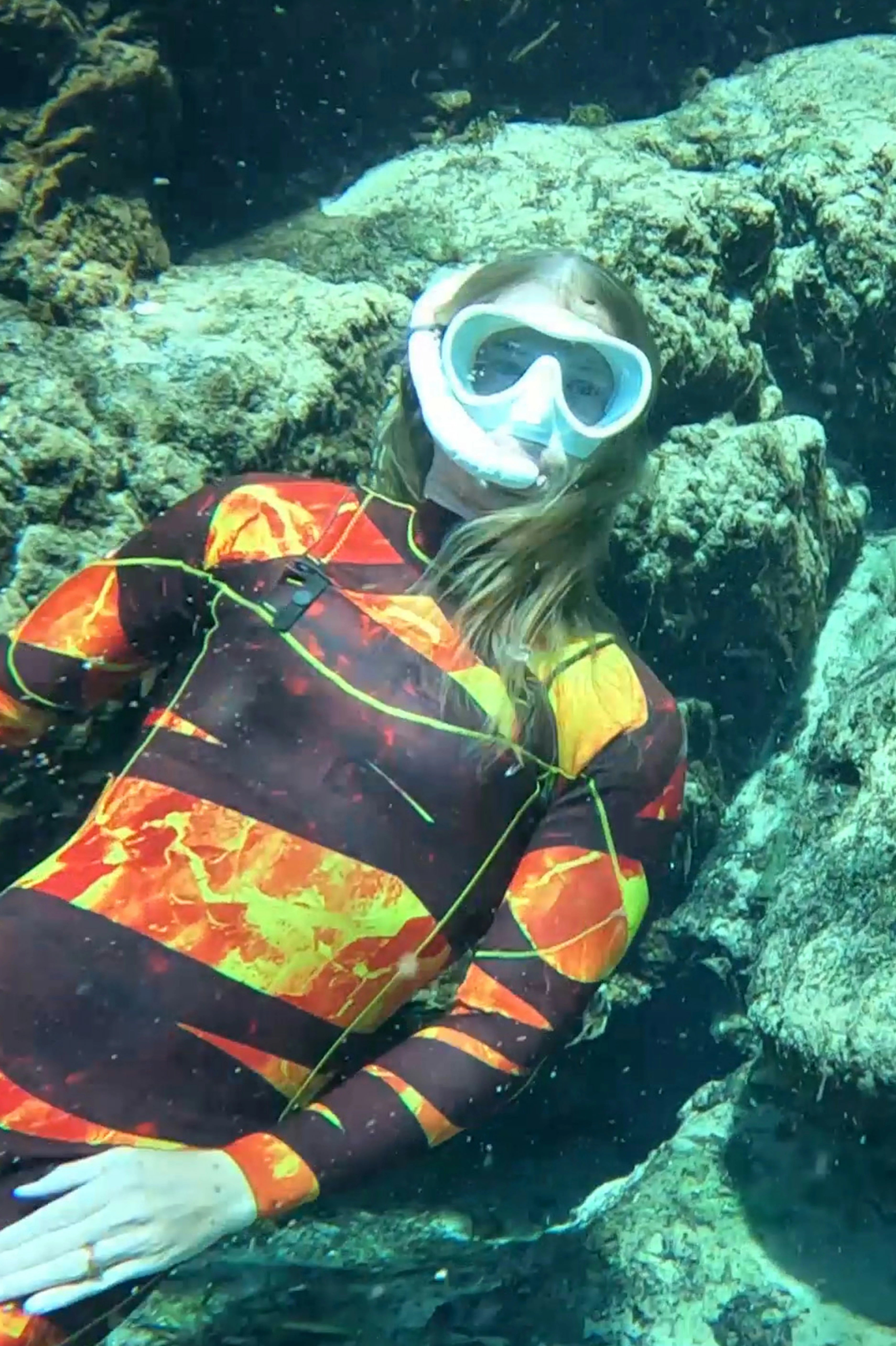 A person underwater wearing a full-face snorkel mask and a long-sleeve Lava dream wetsuit with a bold orange, yellow, and dark pattern, positioned among rocky coral formations.