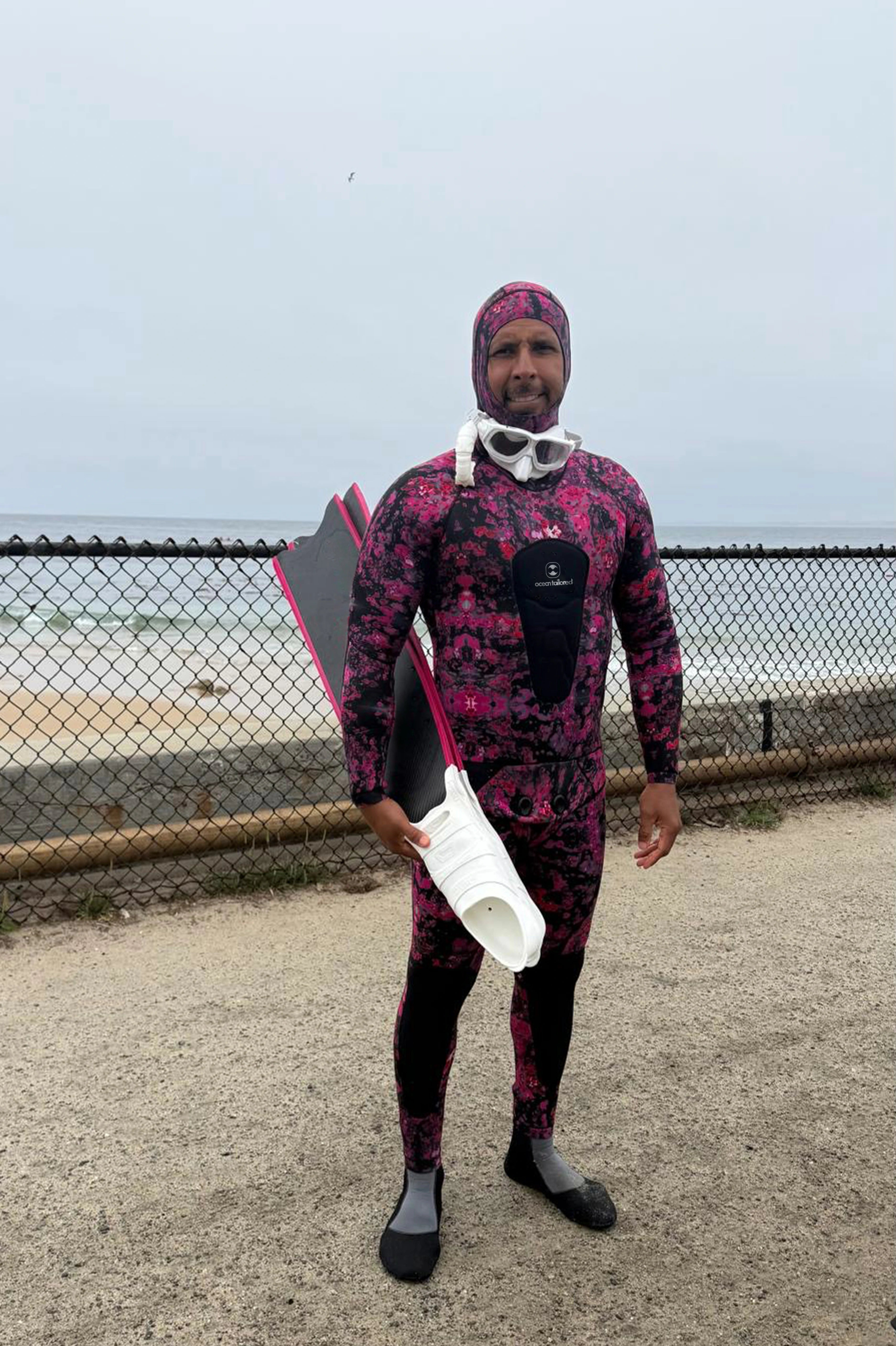 A man wearing a full-body pink and black patterned wetsuit stands on a beach path holding a snorkel and a rolled-up wetsuit or towel, with a chain-link fence and ocean in the background.