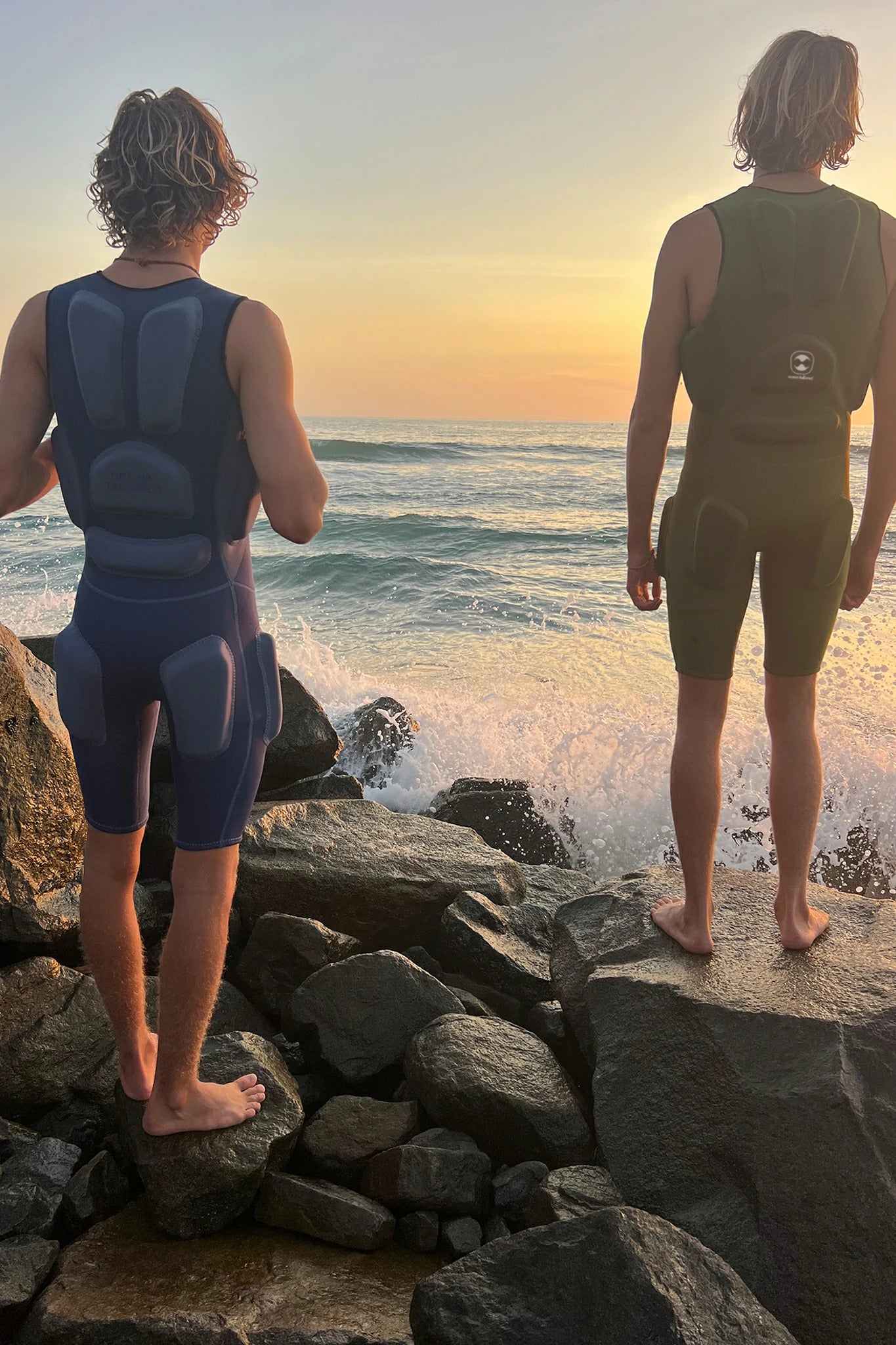 Two people in Hydro Guard Impact Short John wetsuits standing on rocks by the ocean at sunset.