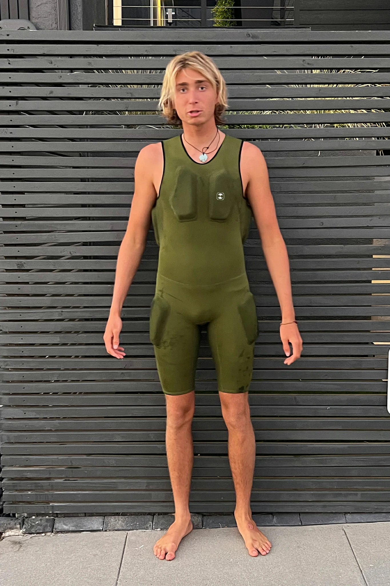 Person wearing a green sleeveless Hydro Guard Impact Short John standing against a wooden wall.