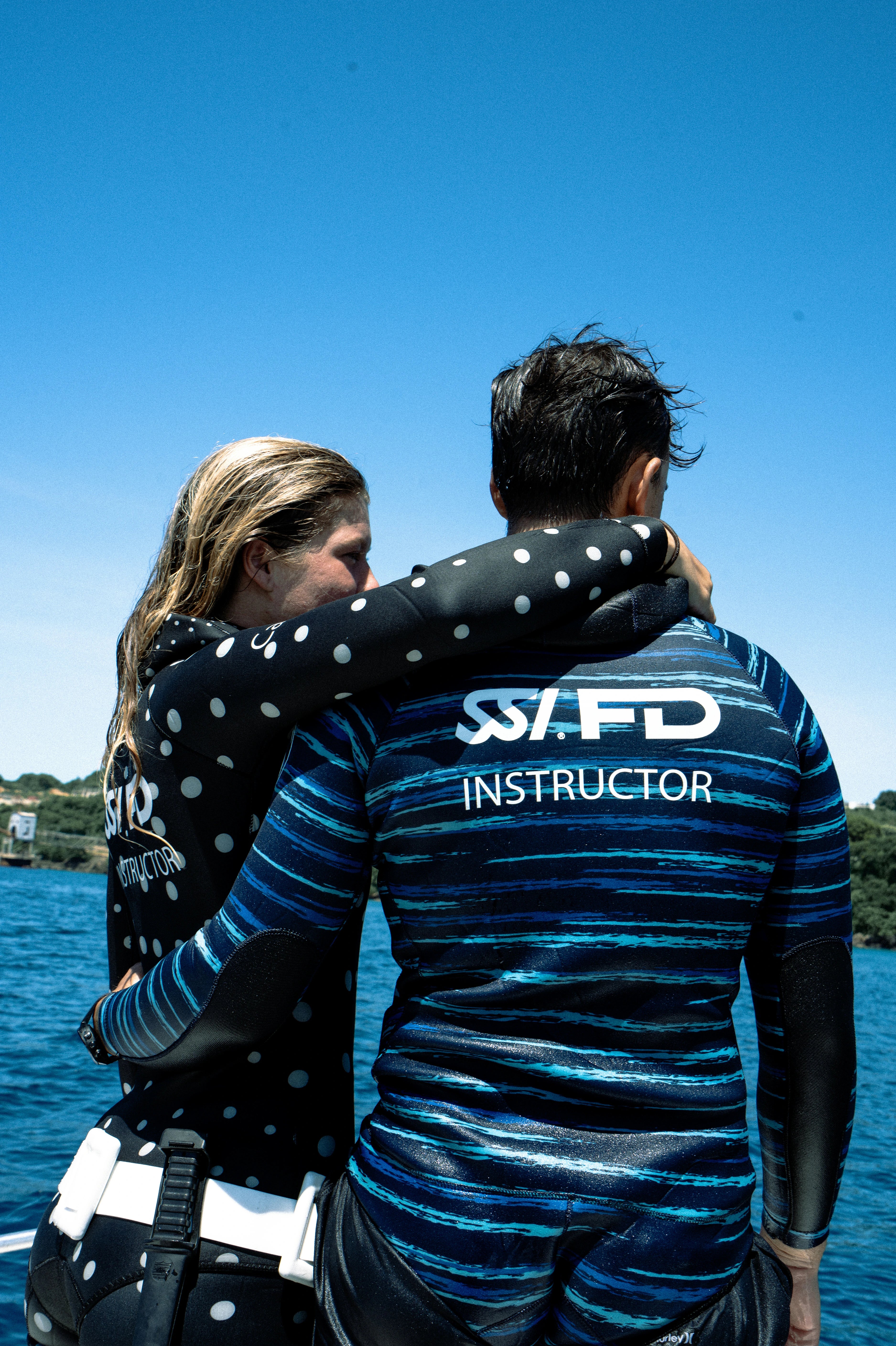 Two people in wetsuits embracing on a boat under a clear blue sky. The person in front wears a dark blue and black striped wetsuit with 'SSFD INSTRUCTOR' printed in white on the back, and the person behind wears a black wetsuit with white polka dots.