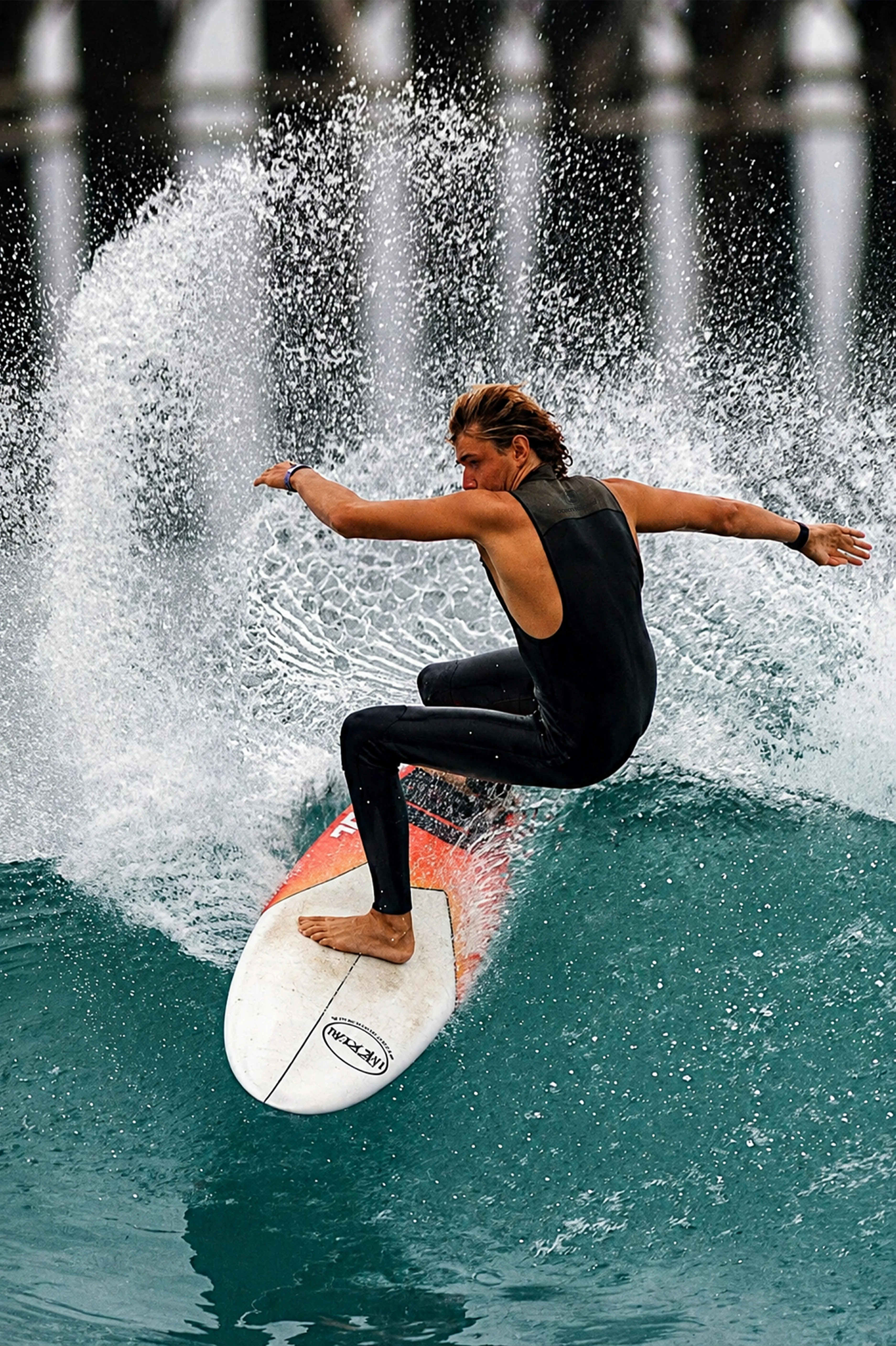 A surfer in a 2mm Long John Wetsuit rides a white and orange surfboard on a turquoise ocean wave, with a large splash of water behind them and a blurred background.