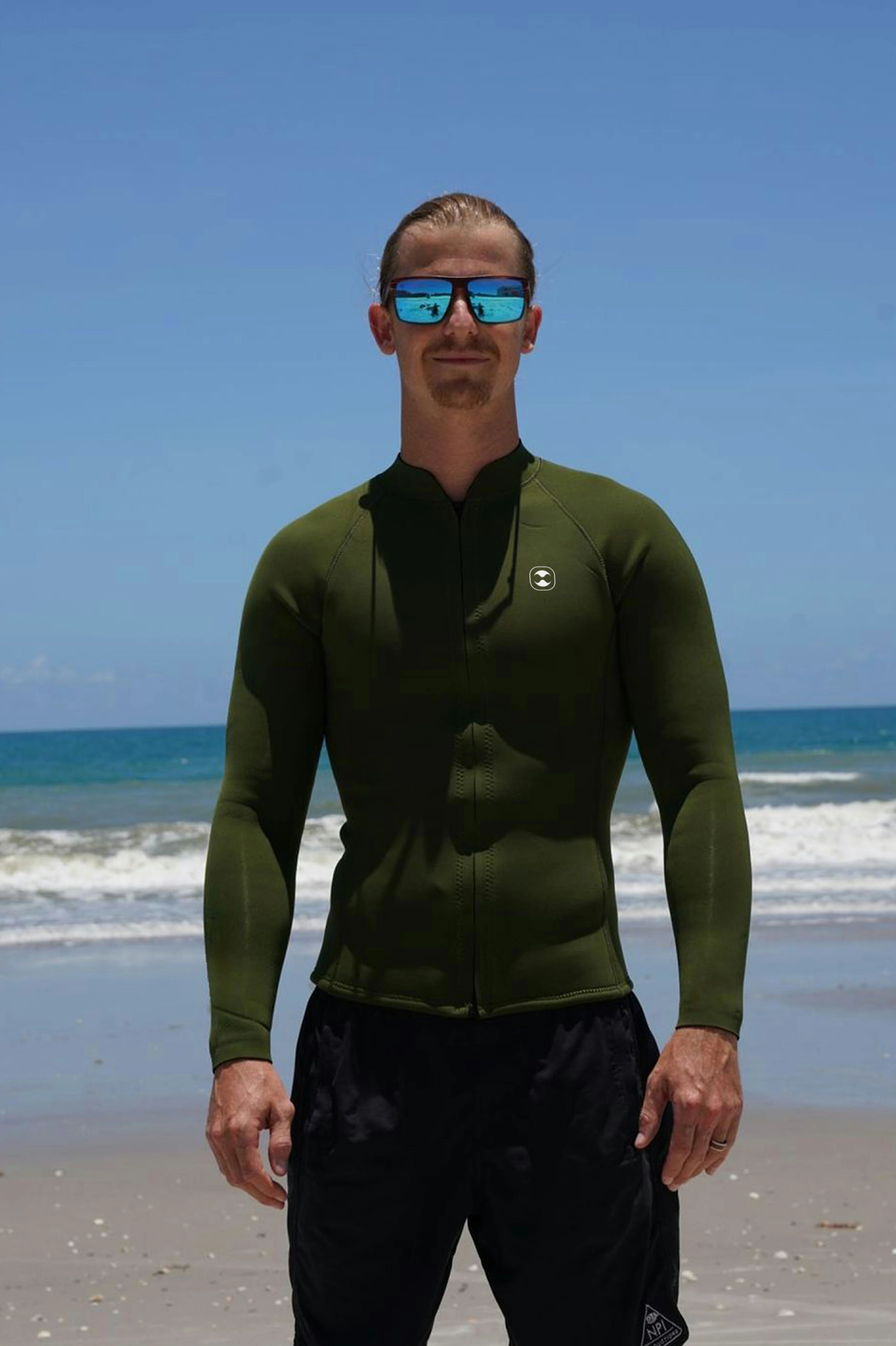 A man standing on a beach wearing a dark olive green long-sleeve 2mm neoprene jacket and black swim trunks, with blue mirrored sunglasses against a background of blue sky and ocean waves.