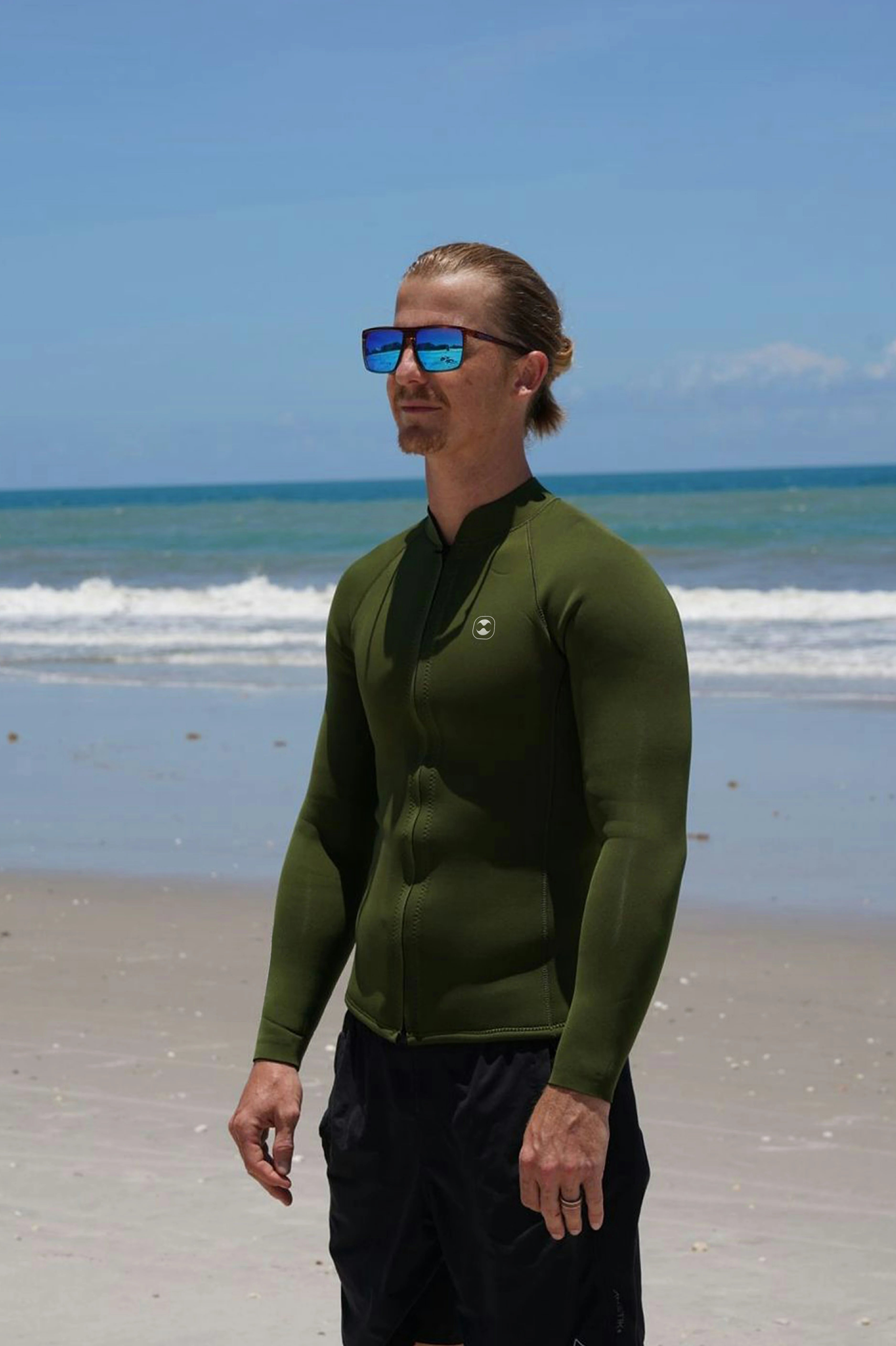 A man wearing an olive green long-sleeve 2mm neoprene jacket and black swim trunks stands on a sandy beach with the ocean and blue sky in the background.