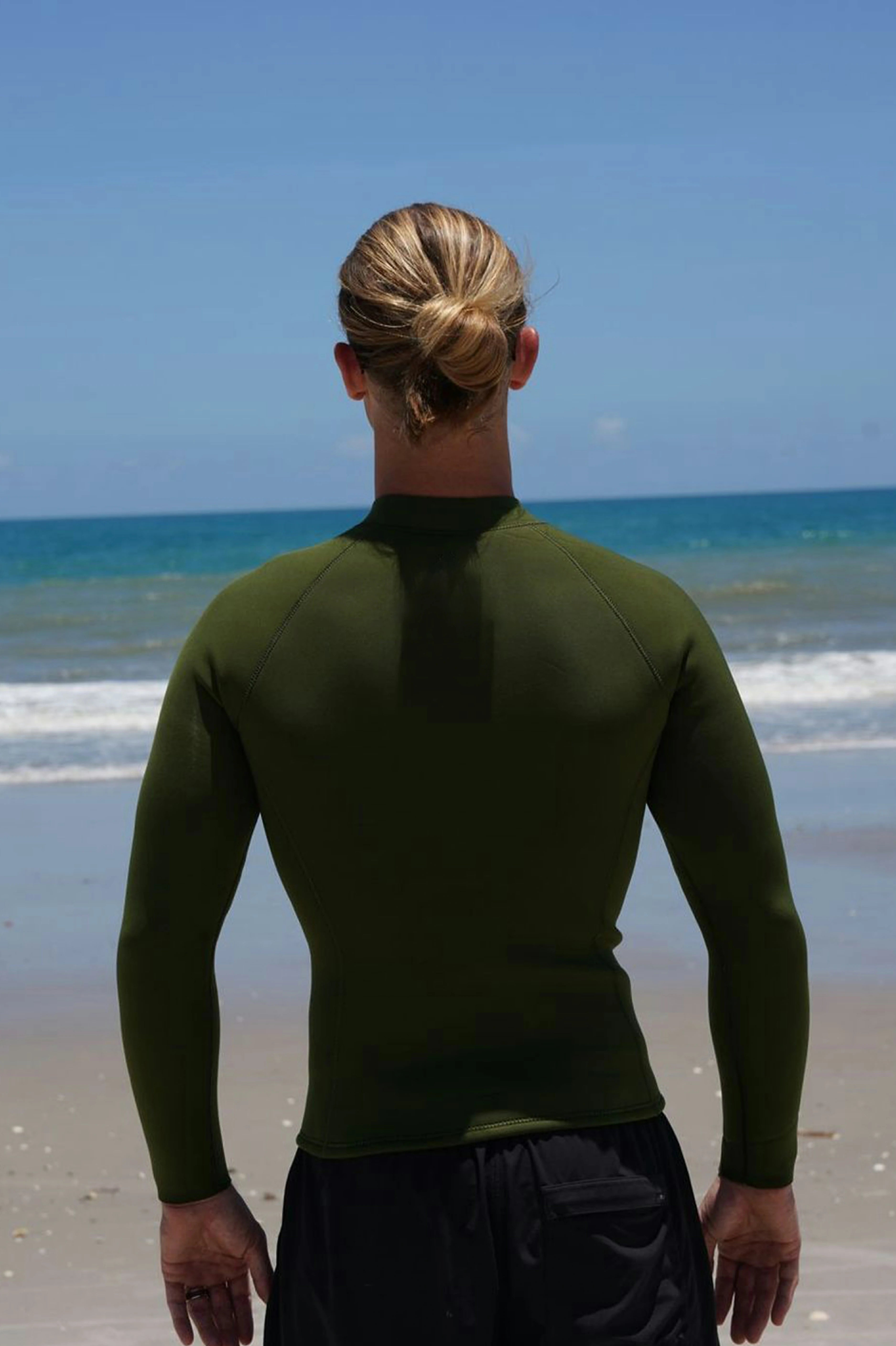 A man standing on a beach with his back to the camera, wearing an olive green long-sleeve wetsuit jacket and black swim trunks, with the ocean and blue sky in the background.