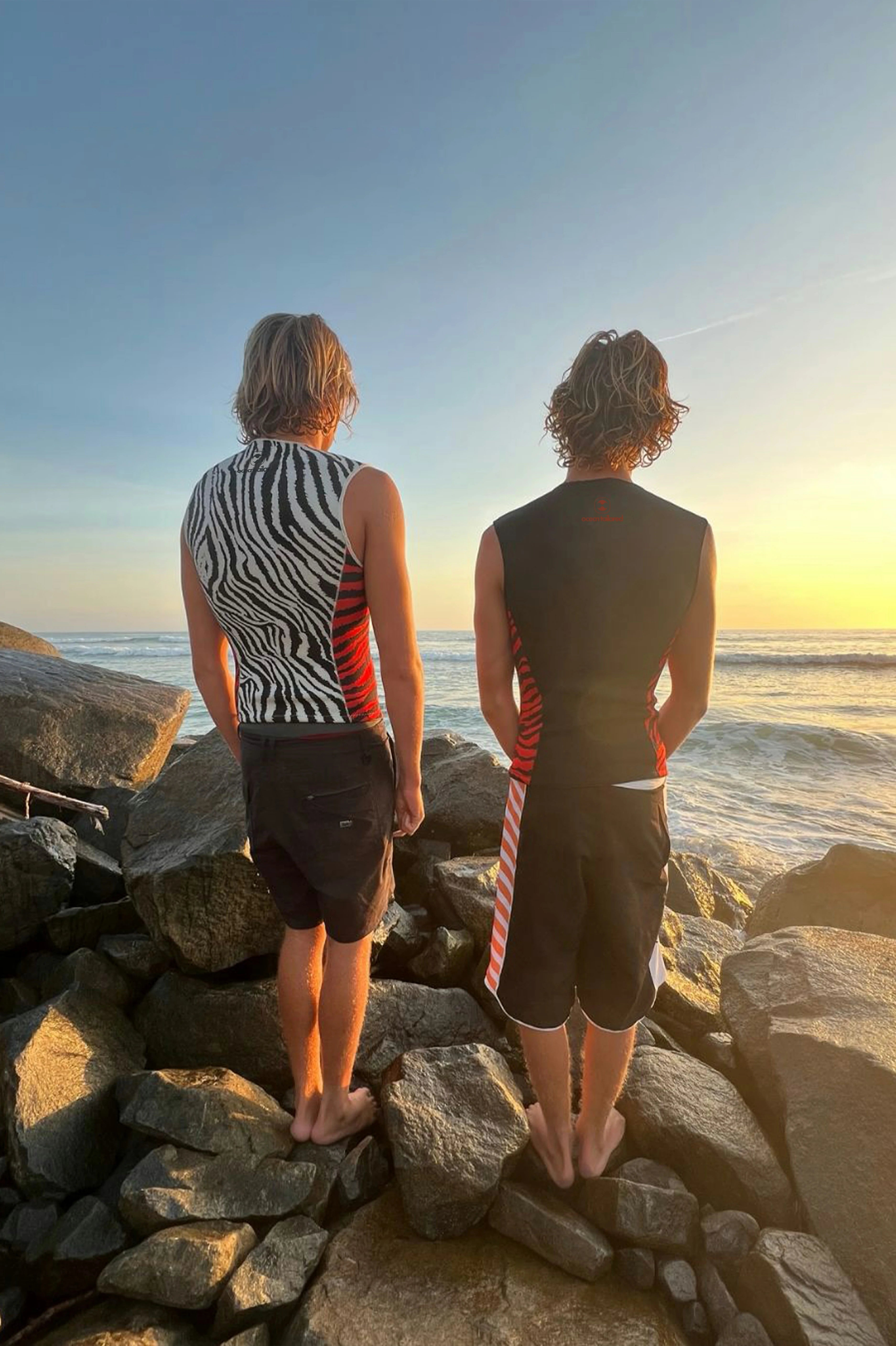 Two people standing on rocks by the ocean at sunset wearing Custom Neoprene Vest.
