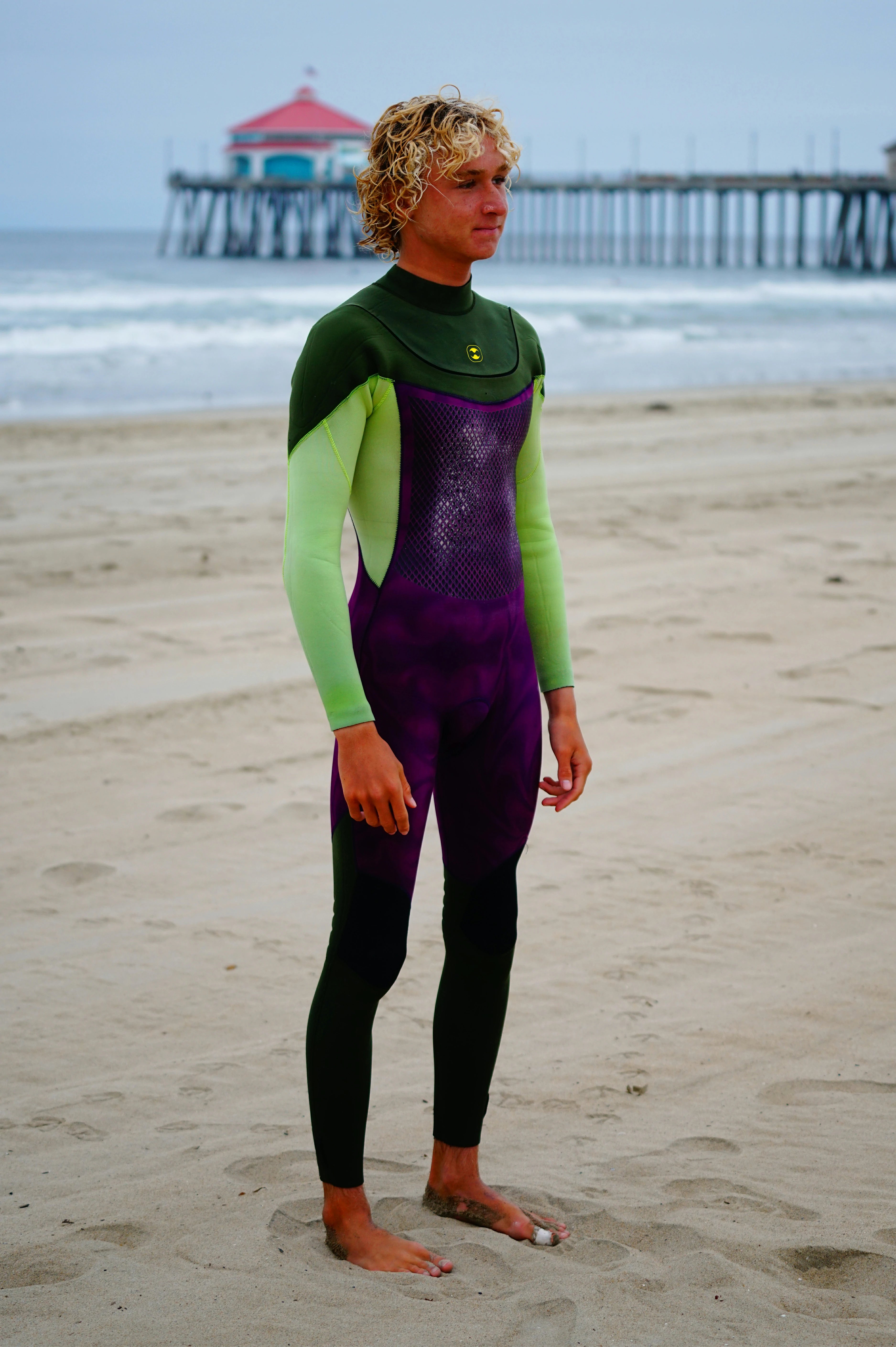 Person wearing a green and purple Flex U-Zip Wetsuit on a beach with a pier in the background