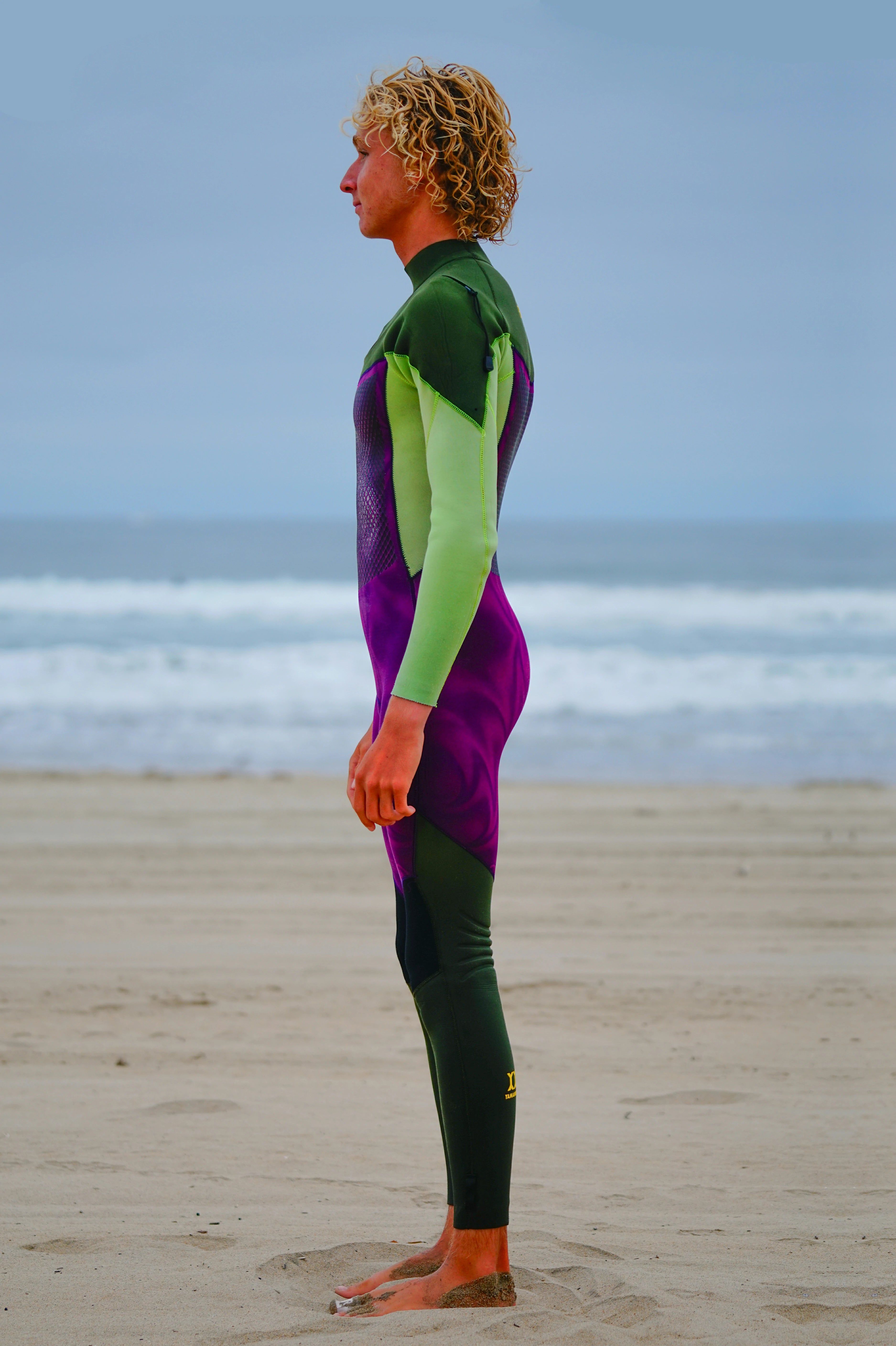 Person wearing a green and purple Flex U-Zip Wetsuit standing on a beach with ocean waves in the background