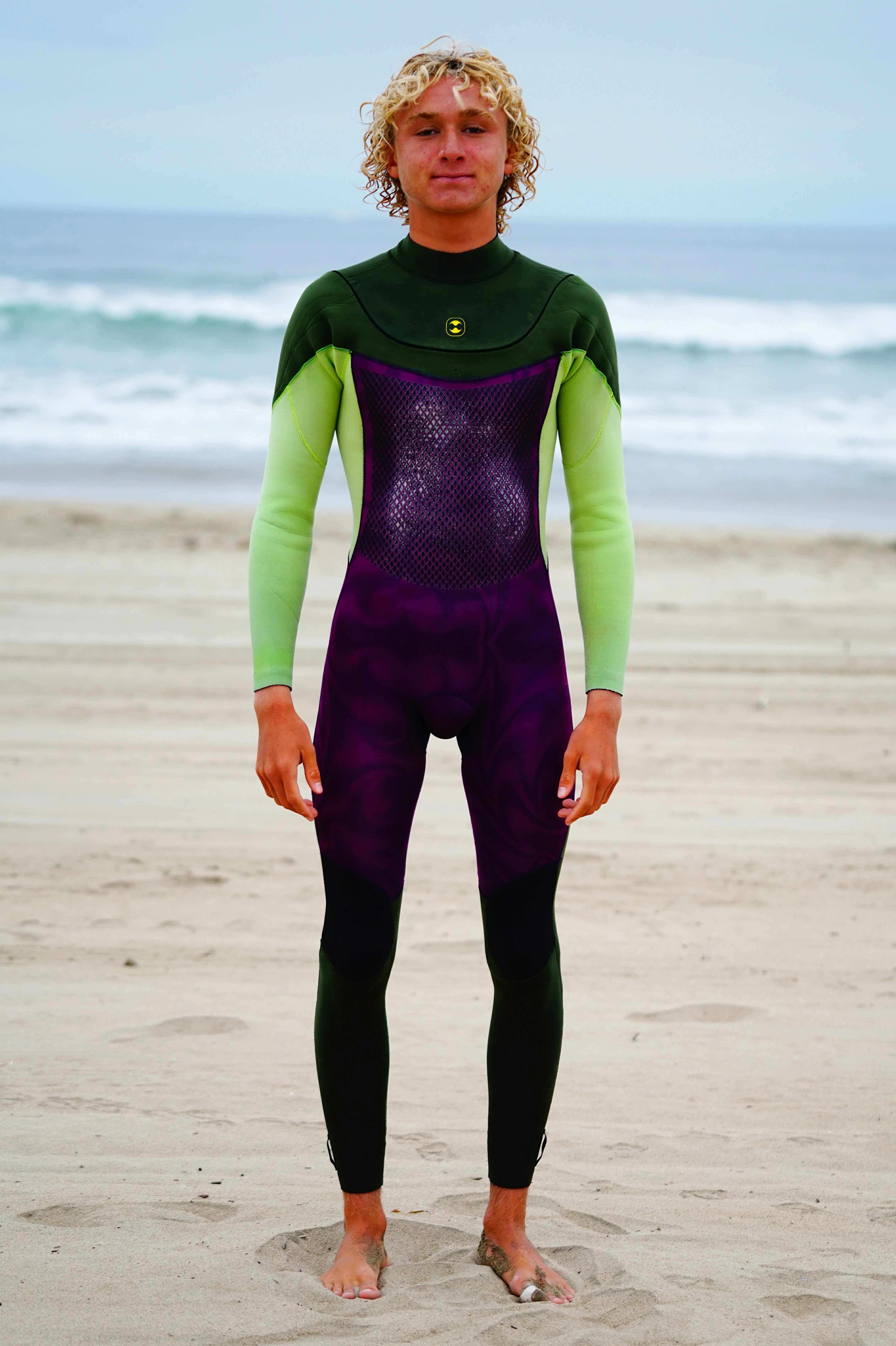 Person wearing a green and purple Flex U-Zip Wetsuit on a beach with ocean waves in the background