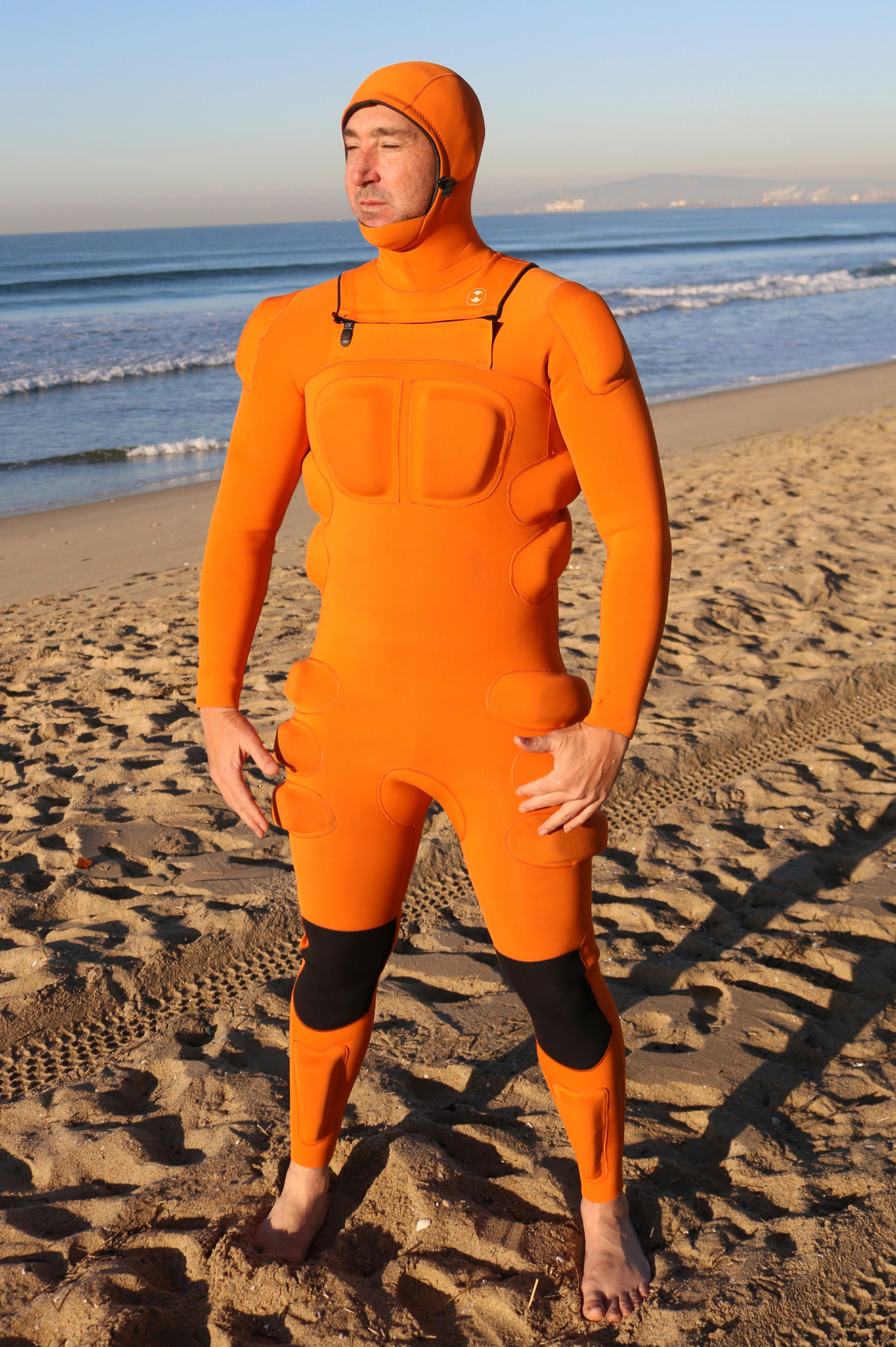 A man wearing a bright orange full-body hydro guard impact wetsuit with black knee panels stands barefoot on a sandy beach with the ocean and sky in the background.