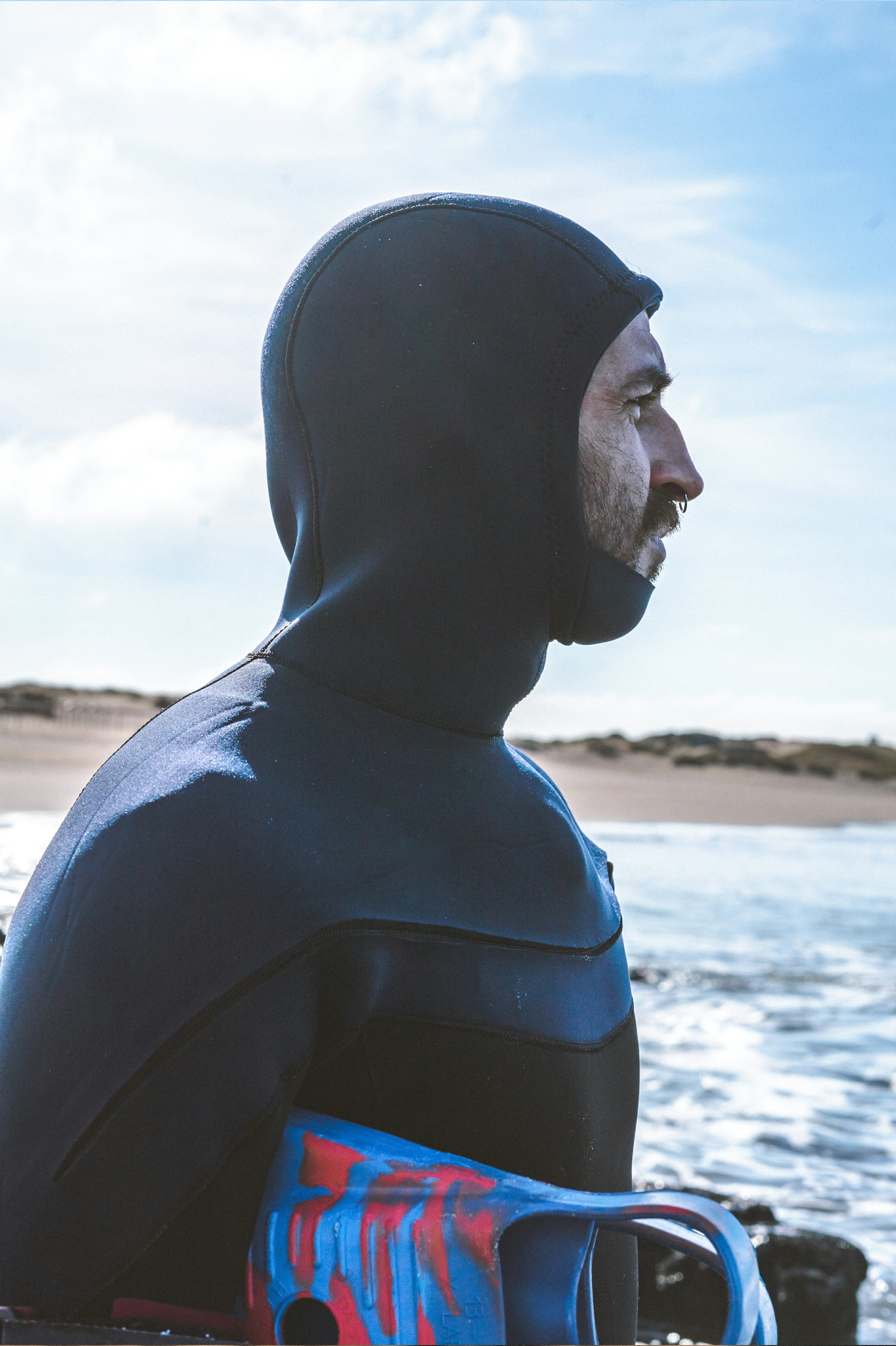 A man in a black full-face wetsuit standing on a beach, holding a blue and red surfboard, with ocean water and a cloudy sky in the background.