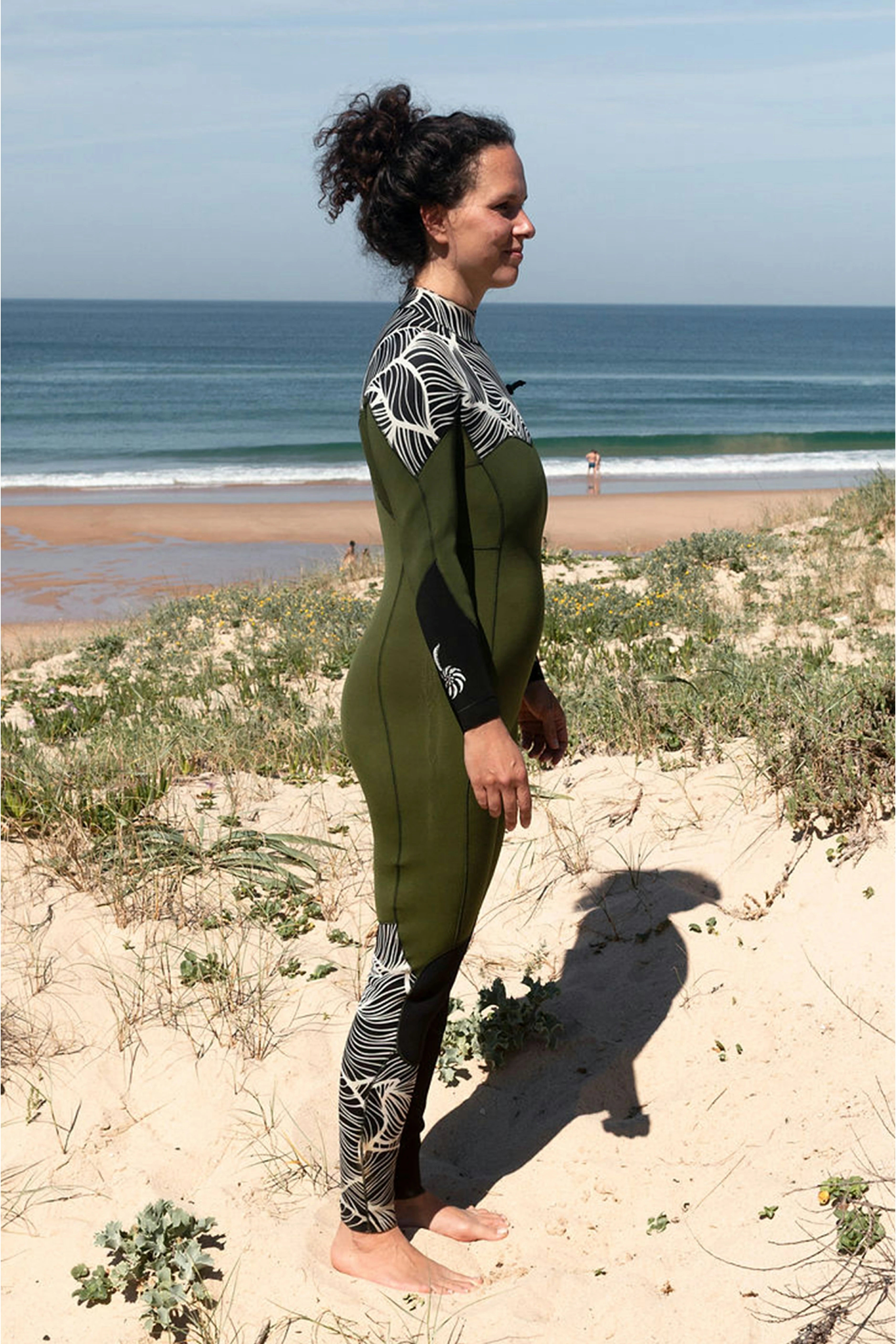 Woman in a green comp zipless wetsuit standing on a sandy beach with ocean in the background