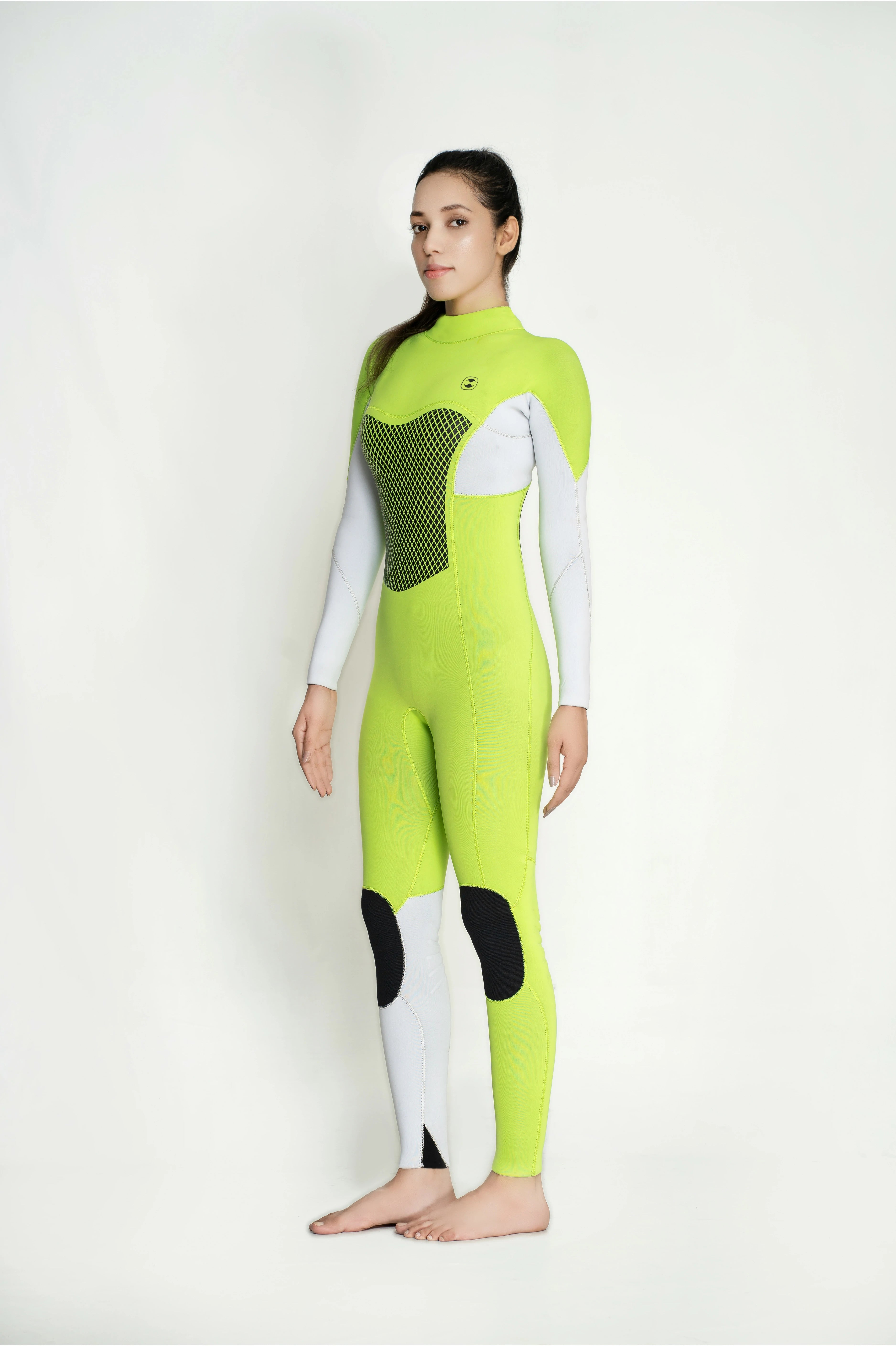 A woman modeling a full-body neon yellow-green classic back zip wetsuit with white sleeves and legs, black knee patches, and a dark mesh chest panel, standing against a plain light background.