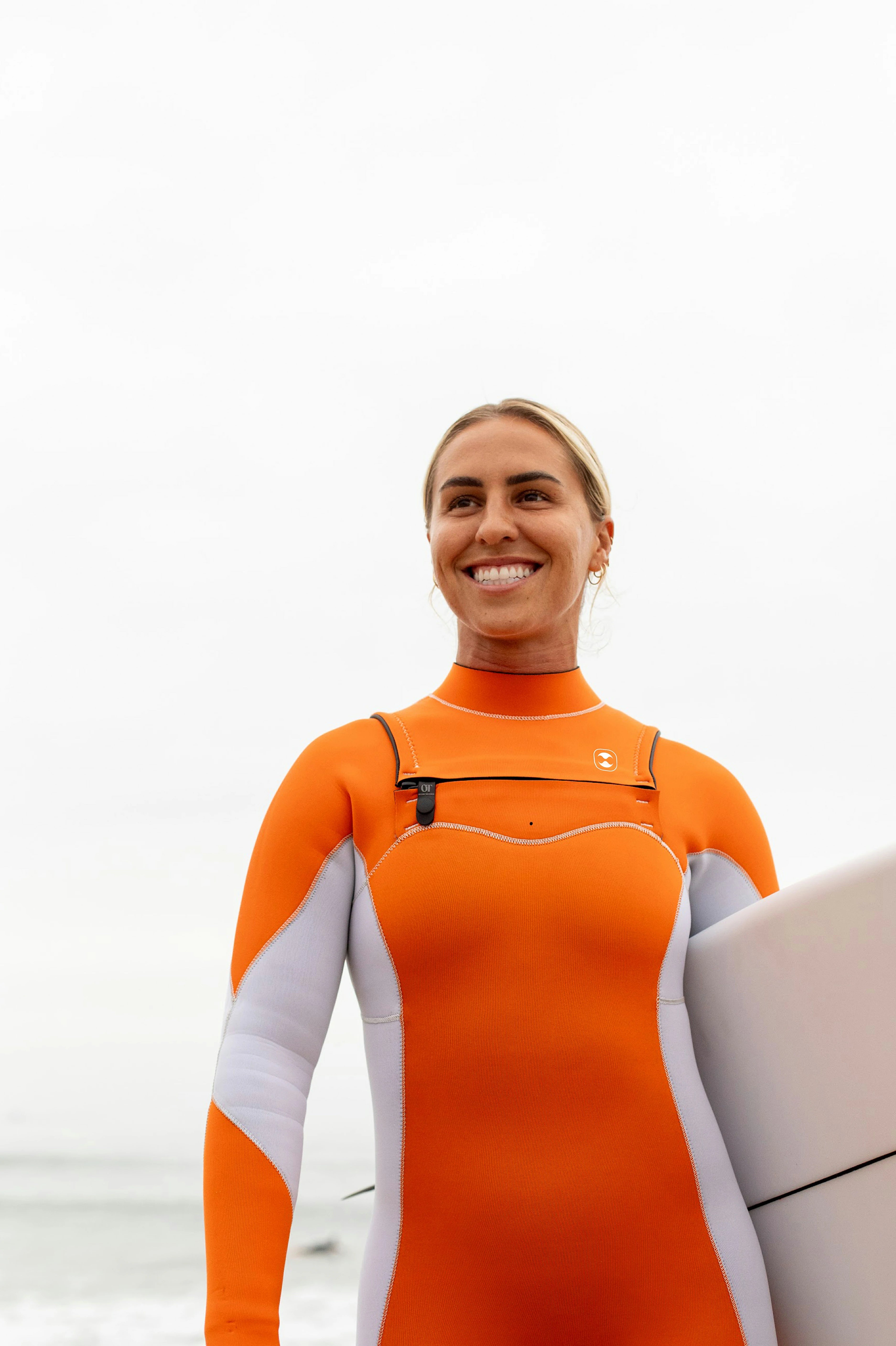 A woman wearing an orange front zip wetsuit holding a surfboard on a beach