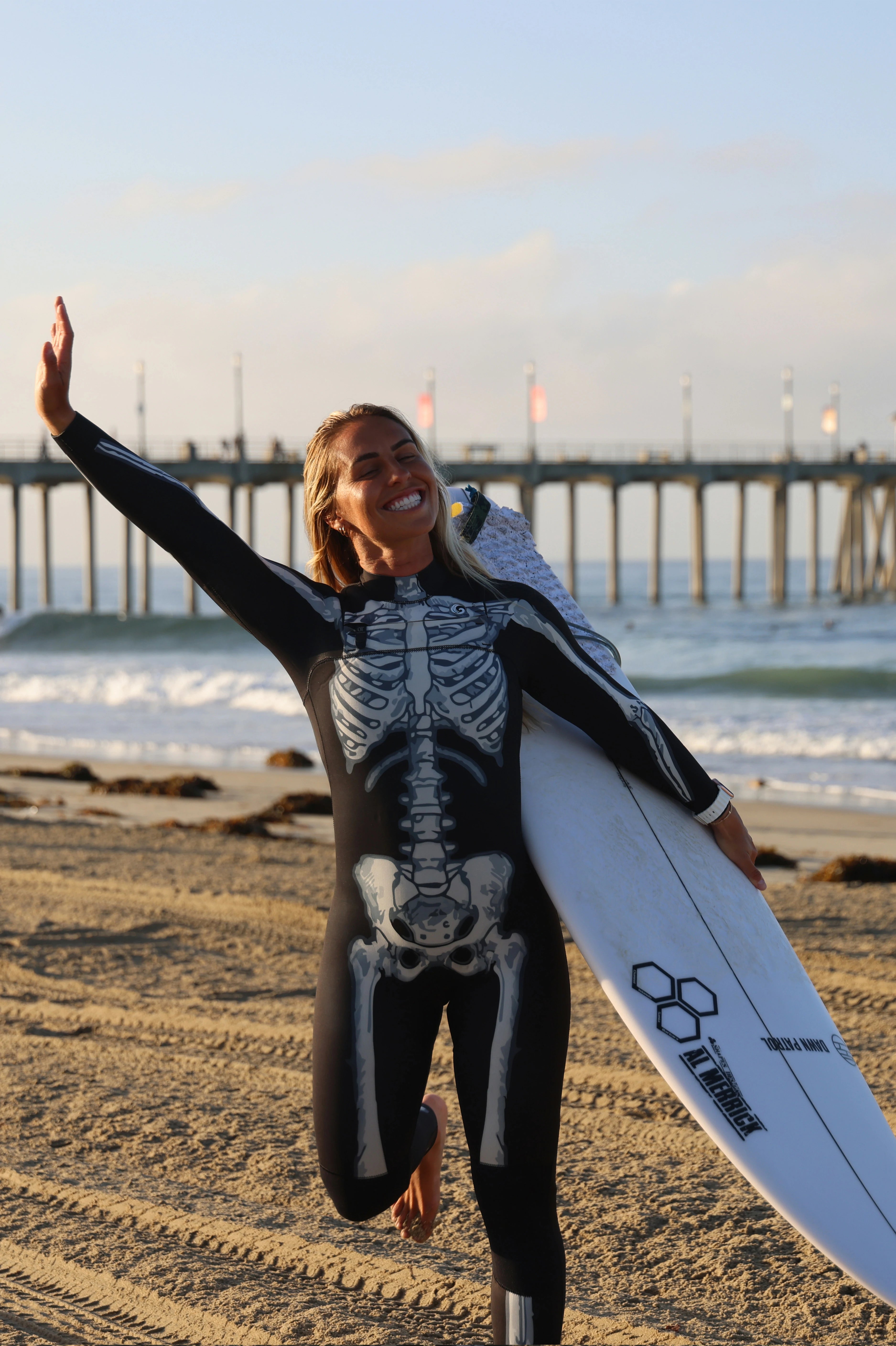 A woman on a sandy beach wearing a black and white skeleton-print wetsuit and carrying a white surfboard, with ocean waves and a pier in the background.