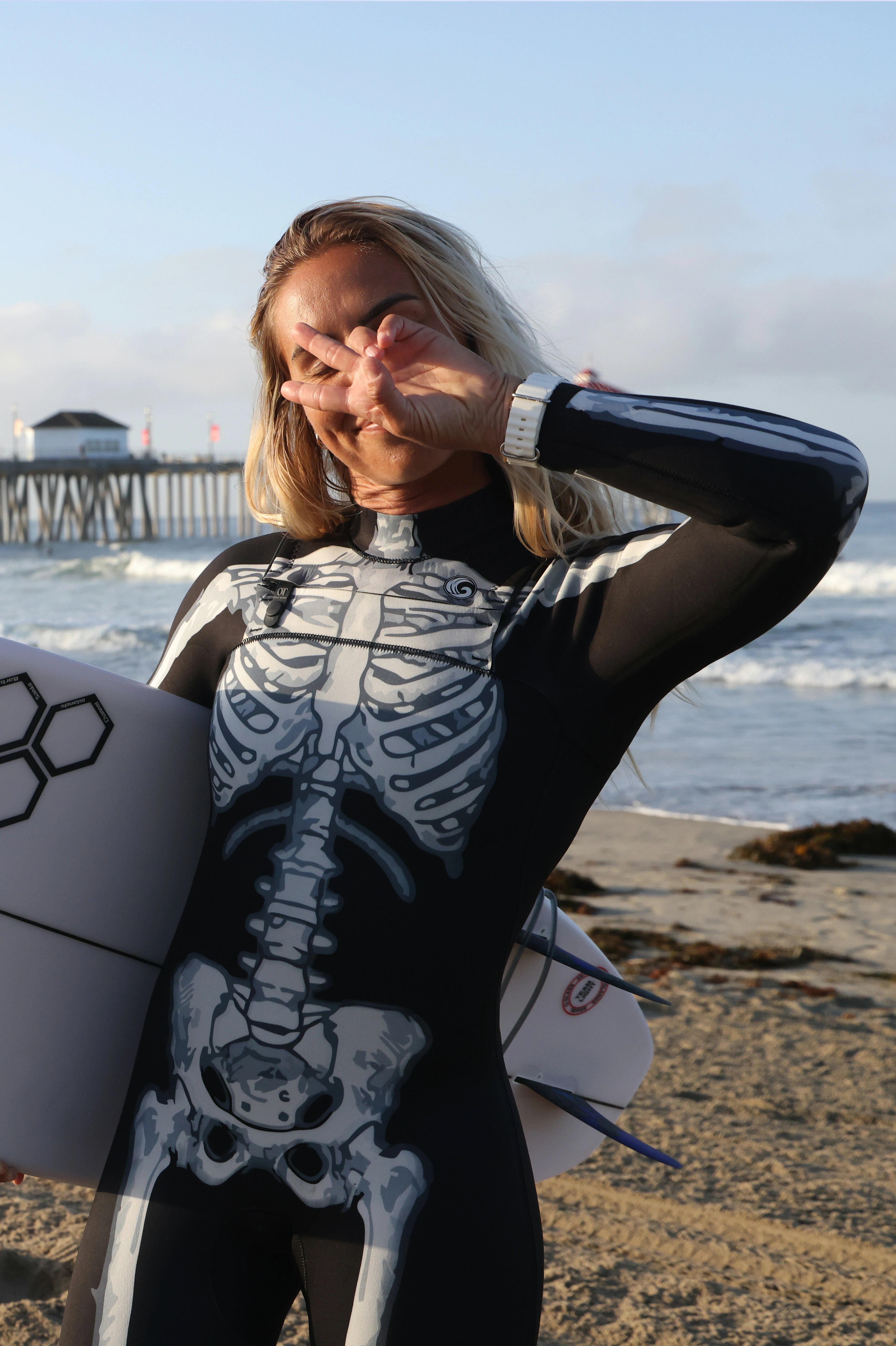 A woman on a beach wearing a black and white skeleton-print wetsuit, holding a white surfboard under her arm. She is posing with her hand over her eyes, with the ocean, pier, and sandy shore in the background.